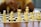A close-up view of a chessboard with white chess pieces prominently positioned, including a king, queen, bishop, knight, and pawns. A person is visible in the background with a blurred yellow shirt, likely contemplating a move.