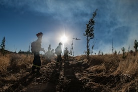 A group of people dressed in firefighting gear walk through a dry, grassy landscape. The sun is shining brightly in the sky, casting long shadows. A helicopter is visible above the scene, suggesting aerial support.