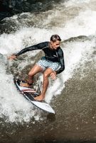 A surfer skillfully rides a wave wearing a black long-sleeve rash guard and blue patterned board shorts. The water splashes energetically around the surfboard, capturing a moment of action and agility.