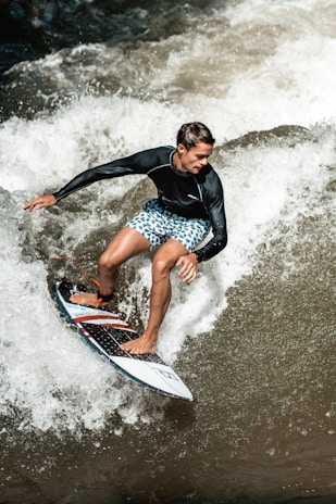 A surfer skillfully rides a wave wearing a black long-sleeve rash guard and blue patterned board shorts. The water splashes energetically around the surfboard, capturing a moment of action and agility.