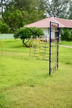 A grassy field featuring a playground with climbing structures made of metal. In the background, a small white building with a red roof is partially visible, surrounded by lush green trees and bushes. The area is enclosed by a light green picket fence.