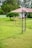A grassy field featuring a playground with climbing structures made of metal. In the background, a small white building with a red roof is partially visible, surrounded by lush green trees and bushes. The area is enclosed by a light green picket fence.