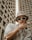 A person wearing a beige bucket hat and large sunglasses is posing with one hand on their face, in front of tall, concrete buildings. Their clothing includes a white shirt with a small heart graphic. The style conveys a sense of urban fashion and the person is accessorized with a chunky gold chain necklace and bracelet.