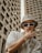 A person wearing a beige bucket hat and large sunglasses is posing with one hand on their face, in front of tall, concrete buildings. Their clothing includes a white shirt with a small heart graphic. The style conveys a sense of urban fashion and the person is accessorized with a chunky gold chain necklace and bracelet.