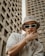 A person wearing a beige bucket hat and large sunglasses is posing with one hand on their face, in front of tall, concrete buildings. Their clothing includes a white shirt with a small heart graphic. The style conveys a sense of urban fashion and the person is accessorized with a chunky gold chain necklace and bracelet.