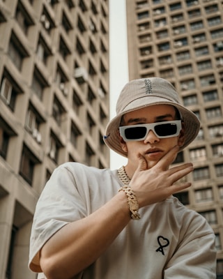 A person wearing a beige bucket hat and large sunglasses is posing with one hand on their face, in front of tall, concrete buildings. Their clothing includes a white shirt with a small heart graphic. The style conveys a sense of urban fashion and the person is accessorized with a chunky gold chain necklace and bracelet.