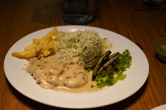 A plate of food featuring a portion of white and green herb rice, a serving of fries, creamy sauced chicken or meat dish, sautéed green beans, and green lettuce leaves. The plate is set on a wooden table with a glass of water in the background.