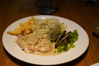 A plate of food featuring a portion of white and green herb rice, a serving of fries, creamy sauced chicken or meat dish, saut&eacute;ed green beans, and green lettuce leaves. The plate is set on a wooden table with a glass of water in the background.