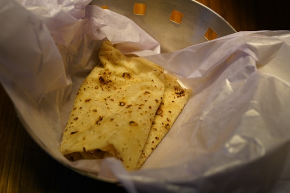 A piece of flatbread with brown spots from baking is wrapped in white paper inside a metallic-looking container.