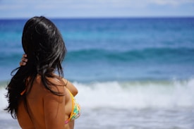 A woman with long dark hair stands at the edge of a beach, looking out towards the blue ocean. She is wearing a colorful bikini and has her hand resting on her hair. The sea is calm, with gentle waves approaching the shore under a clear sky.
