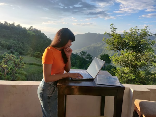 a woman working on a laptop at a table on a balcony, with a mountainous natural vista in the background