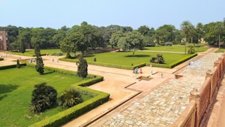 A well-maintained garden with neatly trimmed hedges and a variety of trees, including palm trees. Pathways intersect the lush green lawns, and several small groups of people are strolling or sitting around the area. A stone pathway runs along the right side, bordered by a reddish stone wall, with some architectural features visible in the background.
