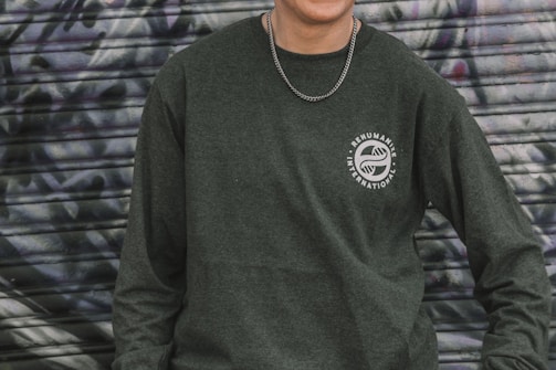 A person is wearing a dark green sweatshirt with the logo 'Rehumanize International' printed on the chest. The background consists of a metal shutter with abstract graffiti patterns in shades of gray and purple. A chain necklace is worn around the neck.