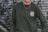 A person is wearing a dark green sweatshirt with the logo 'Rehumanize International' printed on the chest. The background consists of a metal shutter with abstract graffiti patterns in shades of gray and purple. A chain necklace is worn around the neck.