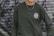 A person is wearing a dark green sweatshirt with the logo 'Rehumanize International' printed on the chest. The background consists of a metal shutter with abstract graffiti patterns in shades of gray and purple. A chain necklace is worn around the neck.