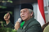 An older individual is speaking into a microphone, wearing a dark hat and a green garment, with a gesture of emphasis. A partially visible flag with red and white stripes is in the background.