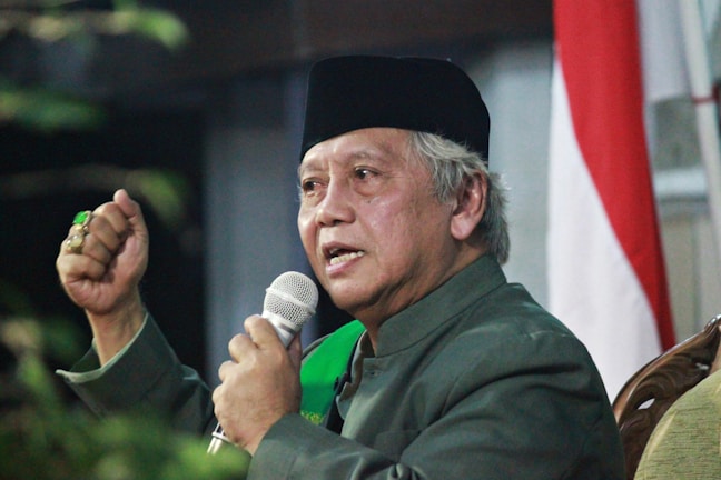 An older individual is speaking into a microphone, wearing a dark hat and a green garment, with a gesture of emphasis. A partially visible flag with red and white stripes is in the background.
