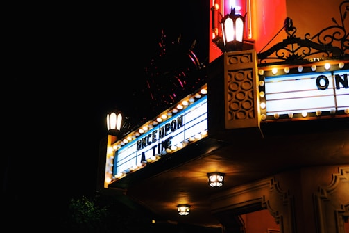 A vintage-style cinema marquee illuminated at night, displaying the words 'ONCE UPON A TIME'. The sign is adorned with classic light bulbs and ornate details. The warm glow of lanterns adds an old-fashioned charm to the scene, contrasting against the dark night sky.