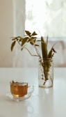 A clear glass cup filled with tea is placed on a white surface next to a glass jar containing green leafy stems. The background is softly lit, creating a calm and serene atmosphere.