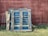 A stack of worn blue wooden pallets rests against a weathered red wooden fence with patches of grass growing at the base.