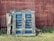 A stack of worn blue wooden pallets rests against a weathered red wooden fence with patches of grass growing at the base.