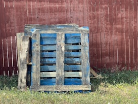 A stack of worn blue wooden pallets rests against a weathered red wooden fence with patches of grass growing at the base.