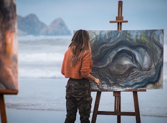 woman in orange hoodie standing on brown wooden seat looking at blue and white ocean waves