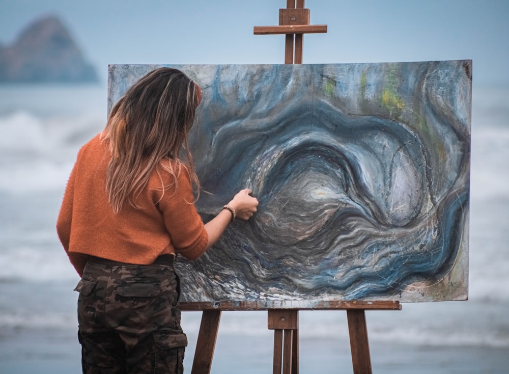 woman in orange hoodie standing in front of blue and white ocean waves painting