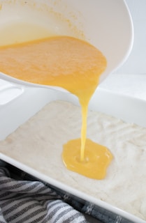 A mixture of what appears to be beaten eggs is being poured from a white bowl into a white rectangular baking dish that contains an unbaked dough base. A gray and white striped kitchen towel is partially visible at the bottom of the image.