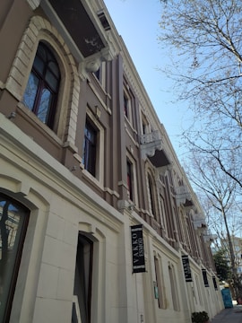 A multi-story building with classical architectural elements, featuring arched windows and decorative columns. The exterior is a mix of beige and white tones with signs displaying the name 'Vakko Esmod.' Leafless trees are visible against a clear blue sky, adding a wintery or early spring atmosphere.
