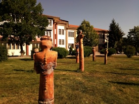 Several wooden sculptures stand on a grassy area in front of a large building with a modern architectural style. The sculptures are tall and simplistic with carved patterns and humanoid features. Surrounding the sculptures are various trees and shrubs, providing a natural setting.