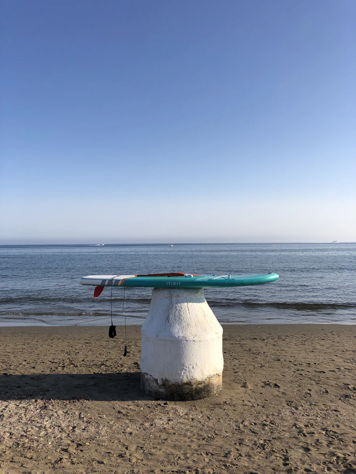 A teal mid-length surfboard resting on a beach plinth with the Pacific Ocean behind