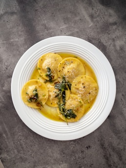 A white plate filled with round ravioli pasta, topped with a light sauce, garnished with green herbs and grated cheese. The ravioli are evenly arranged and the dish sits on a textured gray stone surface.