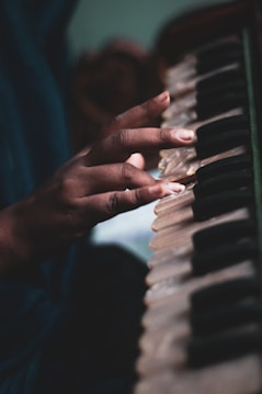 A close-up of a hand playing the keys of a harmonium. The fingers are gently pressing down on the ivory white and black keys, suggesting a moment of musical creation. The background is softly blurred, highlighting the focus on the musician's hand and the instrument.