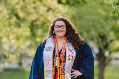 A person is posing outdoors wearing graduation attire, including a cap and gown. They are adorned with various honors cords and a sash with embroidered symbols and the text 'Class of 2021.' The background is a blurred scene of greenery, suggesting a park or garden setting.