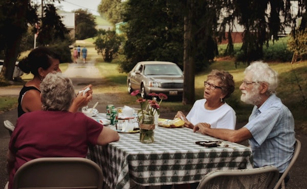 An older couple with glasses chat with two women over a casual meal outside. A checkered tablecloth covers the table.