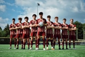 A group of nine young men wearing matching maroon and white striped soccer uniforms stand confidently on a soccer field, with their arms crossed. The grassy field is under a partly cloudy sky, providing a bright, natural backdrop.