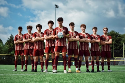 A group of nine young men wearing matching maroon and white striped soccer uniforms stand confidently on a soccer field, with their arms crossed. The grassy field is under a partly cloudy sky, providing a bright, natural backdrop.