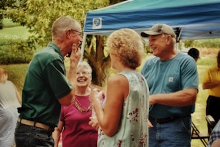 A group of older adults are gathered outdoors under a blue canopy, engaged in a lively and cheerful conversation. There is lush green foliage in the background, contributing to the outdoor setting. The individuals appear relaxed and happy, wearing casual summer clothing.