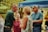 A group of older adults are gathered outdoors under a blue canopy, engaged in a lively and cheerful conversation. There is lush green foliage in the background, contributing to the outdoor setting. The individuals appear relaxed and happy, wearing casual summer clothing.