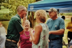 A group of older adults are gathered outdoors under a blue canopy, engaged in a lively and cheerful conversation. There is lush green foliage in the background, contributing to the outdoor setting. The individuals appear relaxed and happy, wearing casual summer clothing.