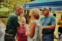 A group of older adults are gathered outdoors under a blue canopy, engaged in a lively and cheerful conversation. There is lush green foliage in the background, contributing to the outdoor setting. The individuals appear relaxed and happy, wearing casual summer clothing.