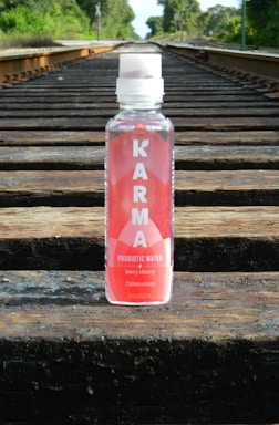A bottle of Karma Probiotic Water with a berry cherry flavor is placed on wooden railroad tracks. The background features lush green foliage alongside the tracks, giving a sense of depth and perspective.