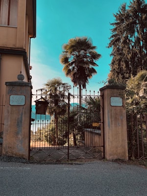 The image depicts a gated entrance labeled with 'Villa Giulia' on stone pillars, surrounded by lush palm trees and greenery. In the background, there is a glimpse of a tranquil body of water, possibly a lake, under a clear, vibrant blue sky.