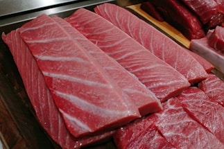 Several thick, rectangular slices of raw fish, likely tuna, displayed on a wooden board. The flesh has a rich, pinkish-red color with visible marbling and a moist, fresh appearance.