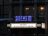 A building facade with a prominently lit marquee displaying the text 'GOT VAX? BE A LOT COOLER IF YOU DID.' in large capital letters. Above the marquee, the word 'ANTHEM' is illuminated in blue neon lights. The building seems to be an entertainment venue or theater with large windows and lighting fixtures visible inside.