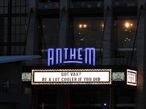 A building facade with a prominently lit marquee displaying the text 'GOT VAX? BE A LOT COOLER IF YOU DID.' in large capital letters. Above the marquee, the word 'ANTHEM' is illuminated in blue neon lights. The building seems to be an entertainment venue or theater with large windows and lighting fixtures visible inside.