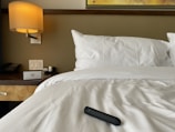 A neatly made bed with white sheets and a black cylindrical object resting on top. A wooden bedside table is visible, featuring a telephone, a light switch panel, and a lamp with a warm glow. A section of a framed wall art piece can be seen above the bed.