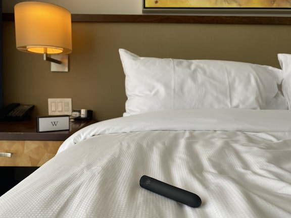 A neatly made bed with white sheets and a black cylindrical object resting on top. A wooden bedside table is visible, featuring a telephone, a light switch panel, and a lamp with a warm glow. A section of a framed wall art piece can be seen above the bed.