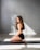 A woman with long wavy hair is kneeling on a yoga mat in a well-lit room. She is wearing a black sports bra and shorts, and her pose suggests she is engaged in a yoga or meditation practice. The room has a minimalistic design with light gray walls and large windows casting shadows on the floor.